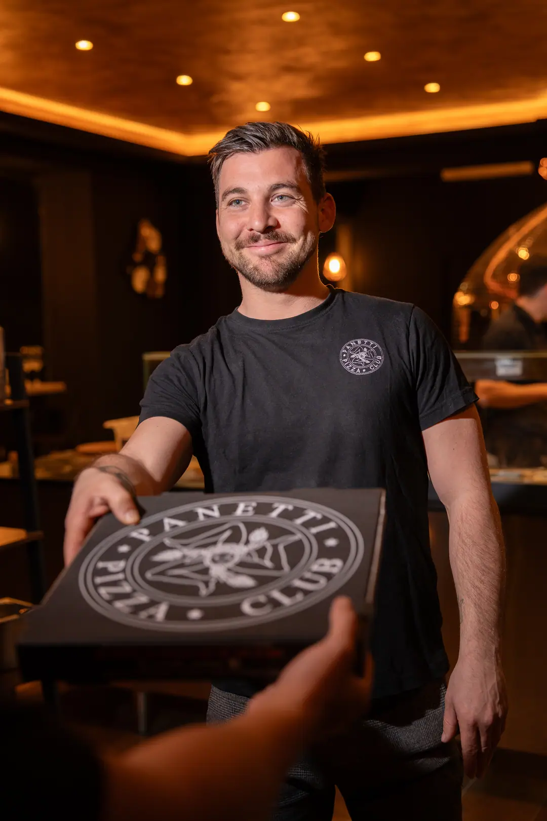 Le pizzaiolo, souriant, tend une boîte Panetti Pizza Club